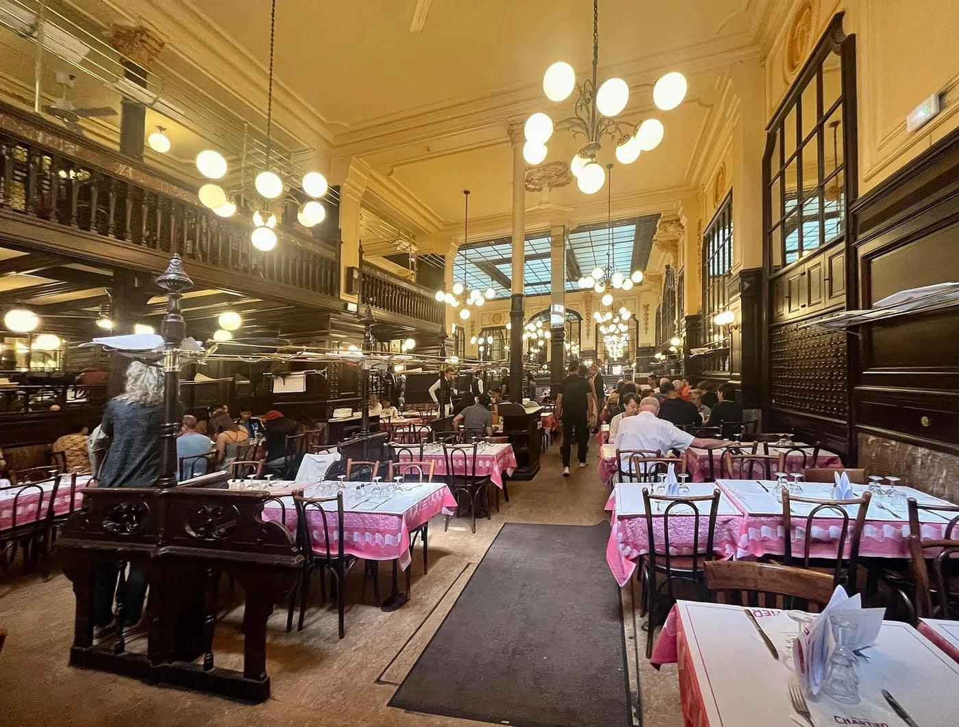 The main dining room of Bouillon Chartier, with tables laid in pink cloths, bentwood chairs, globe chandeliers, a wooden gallery and a glazed roof