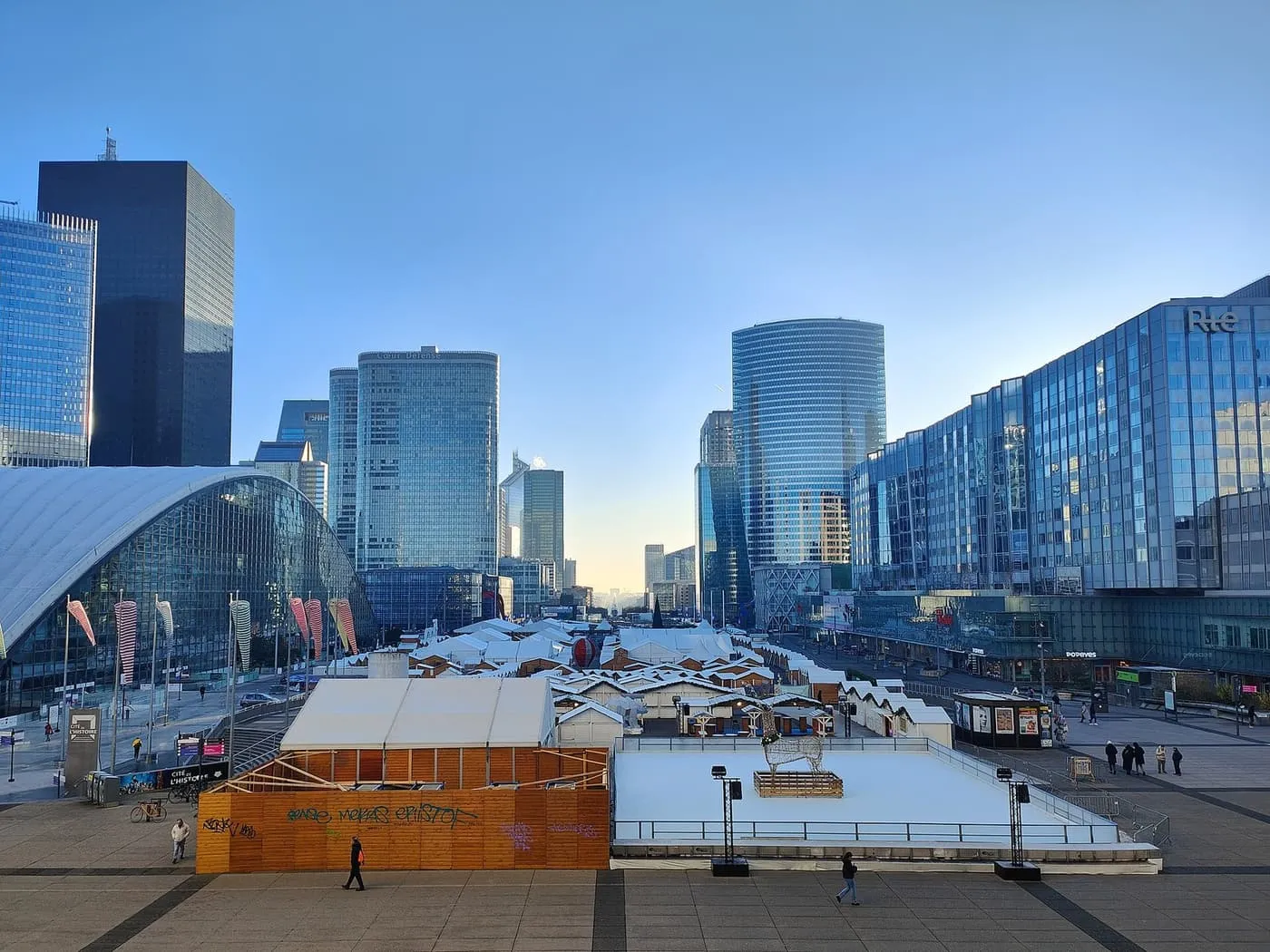 White market chalets and an ice rink on the paved parvis of La Défense, between glass office towers under a clear blue sky